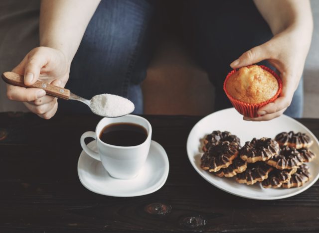 mulher derramando açúcar no café, segurando o muffin acima do prato de biscoitos